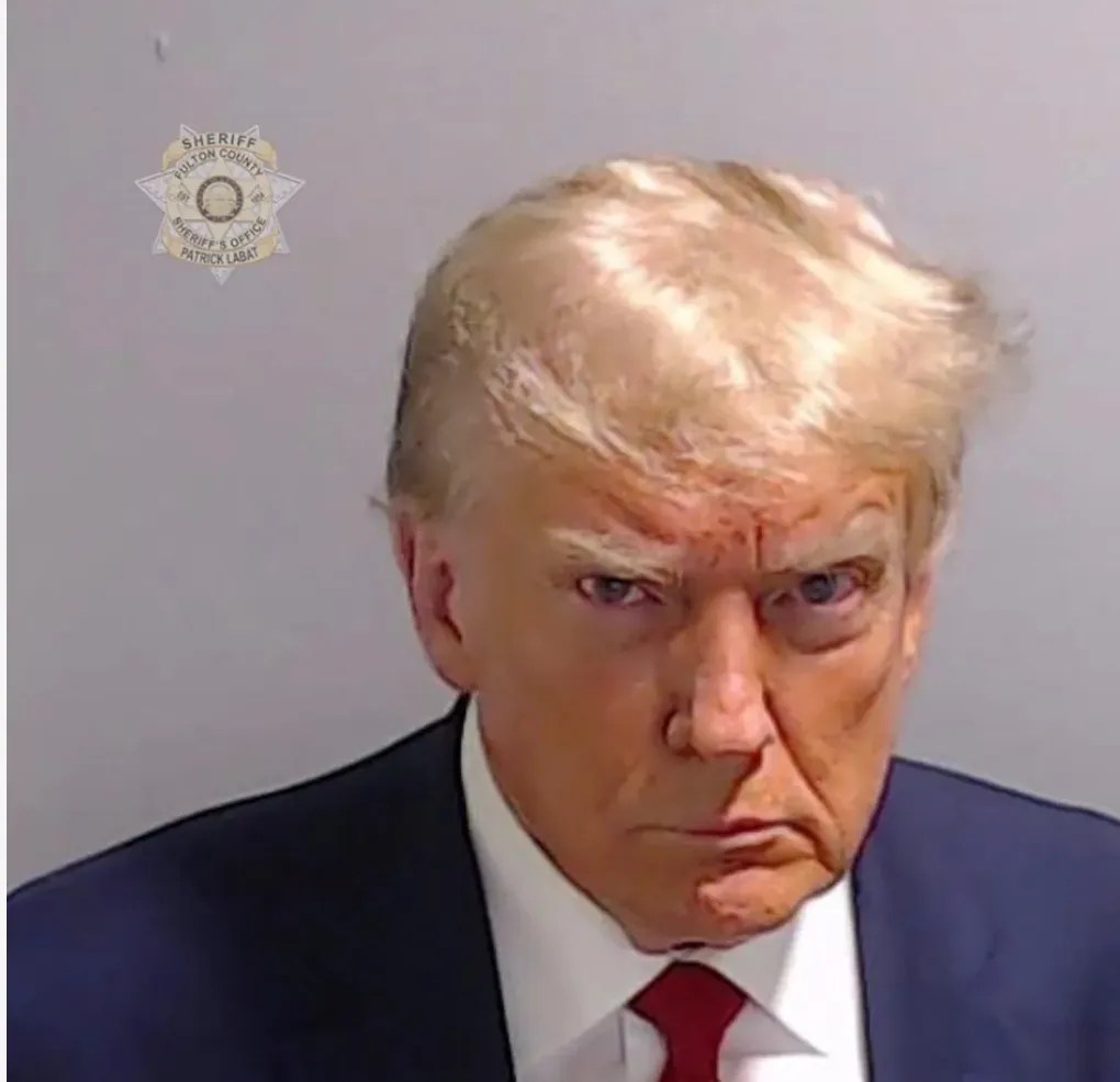 The image shows a person in a suit with a serious expression. The person's hair appears yellow or light blonde hair. He is...